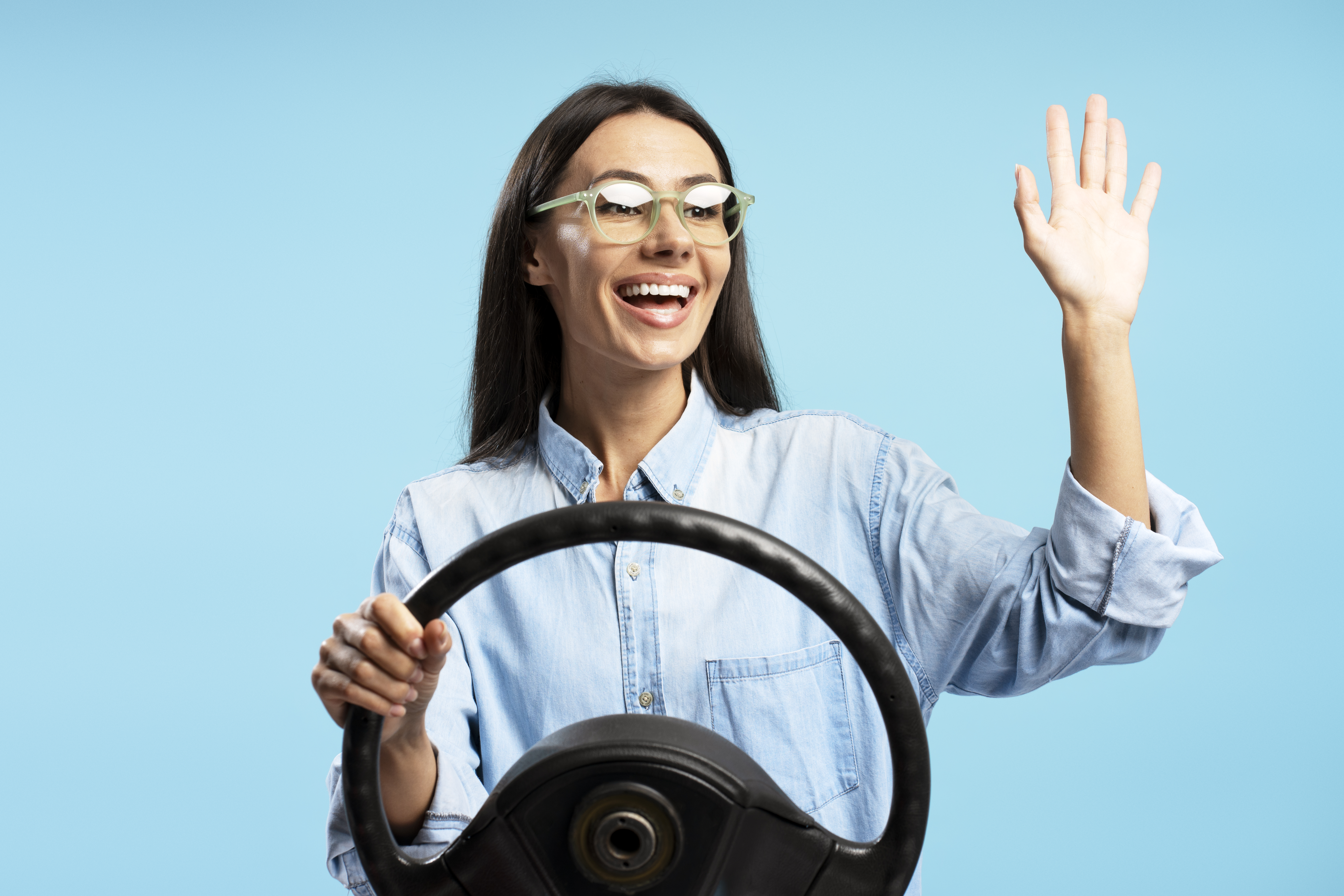 Portrait of smiling cheerful Hispanic woman holding a steering wheel, waving, and smiling against a vibrant blue backdrop, conveying a sense of joy and freedom related to driving. Driving concept